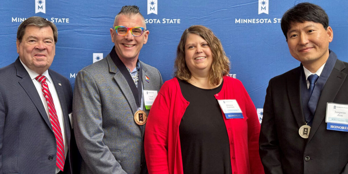 Three men and one woman smile in front of a blue Minnesota State background