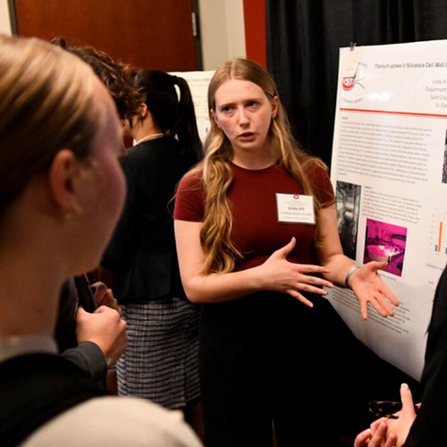 A woman with long blonde hair and a nametag points to a poster presentation